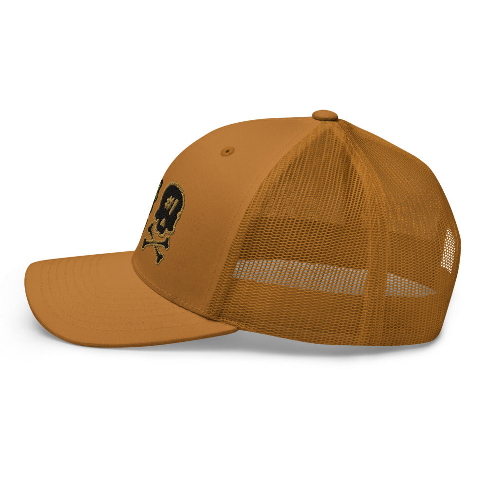 Side view of the caramel SBD #1 trucker cap with the visor pointing left, showing the mesh panels and skull emblem near the front.